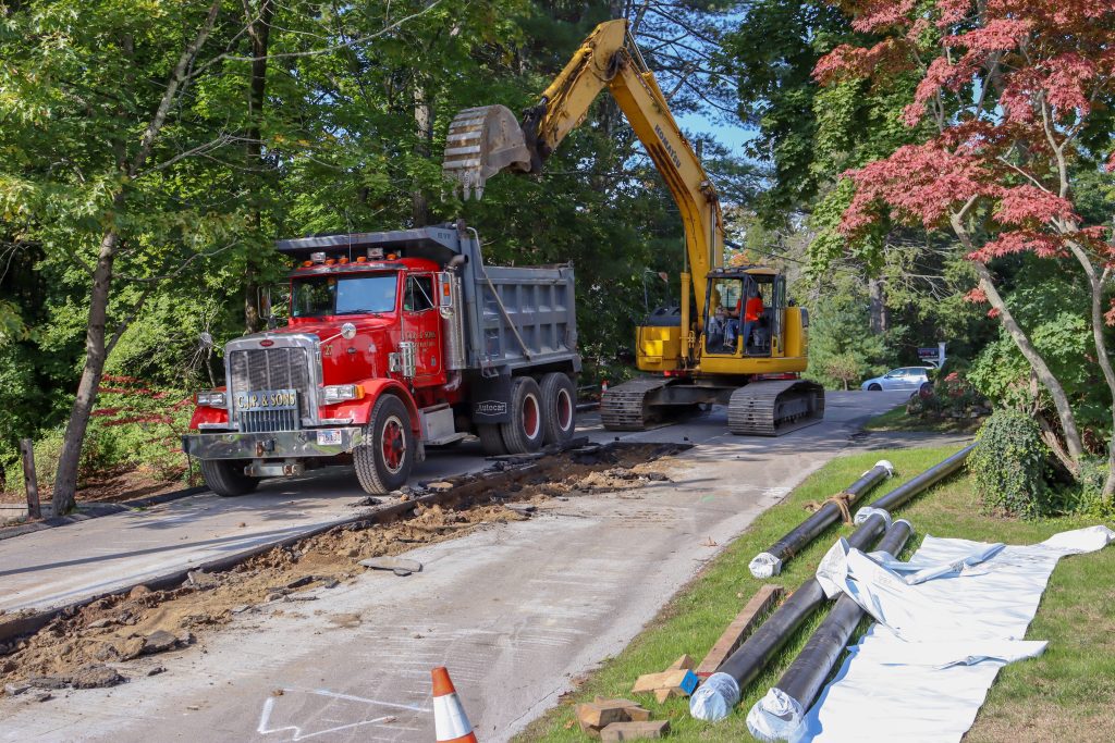 Image of new water main waiting to be installed. An excavator is digging in the road for new water main,.